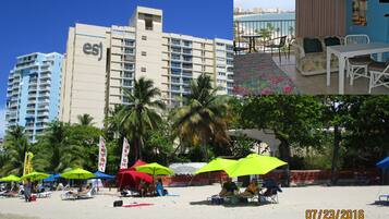 A hotel with a balcony overlooking a beach, a dining area with a view, and a beach area with umbrellas and lounge chairs.