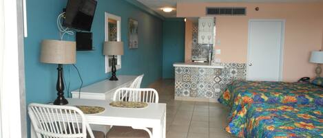 A compact hotel room with a dining area, a kitchenette, a bed with a colorful floral bedspread, and a TV mounted on the wall.