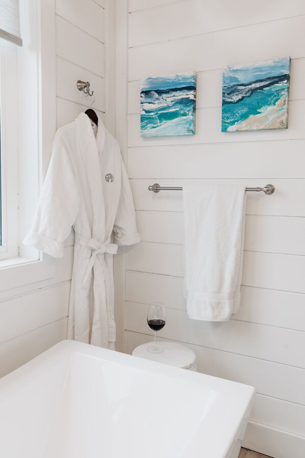A bathroom with a white bathtub, a white robe hanging on a rack, and two paintings of the ocean on the wall.