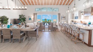 Un vaste espace de vie comprenant un coin repas avec une table, des chaises en osier, une cuisine dotée d’un grand îlot central et une vue sur l’océan grâce à de grandes fenêtres.