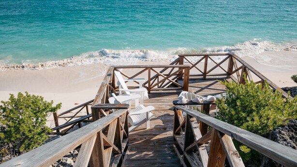 Une terrasse en bois mène à des chaises de plage et à une petite table, offrant une vue sur une plage de sable et une mer turquoise.
