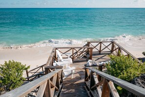 A wooden deck leading to beach chairs and a small table, overlooking a sandy beach and turquoise sea.