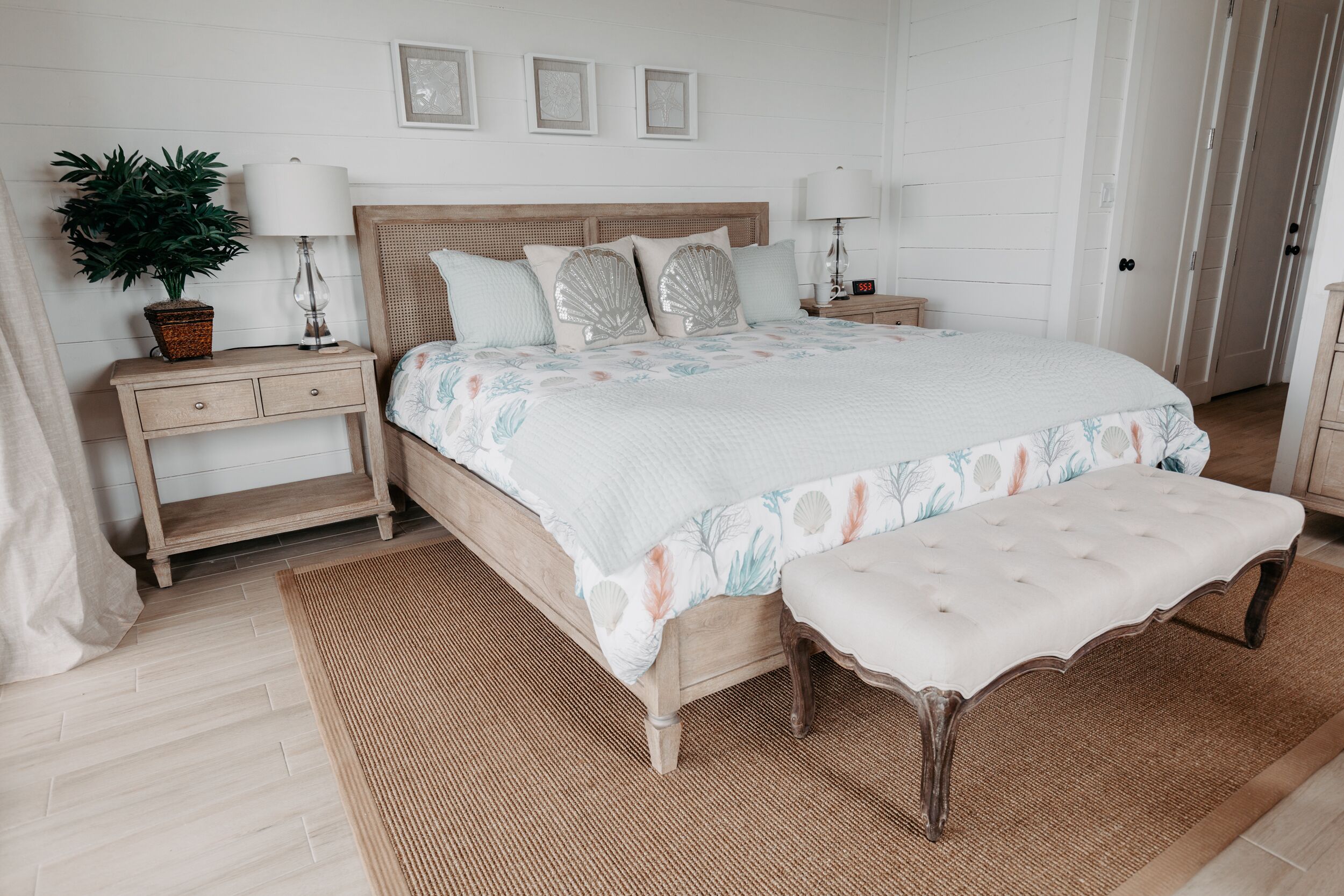 Quarto com uma cama grande, duas mesinhas de cabeceira, um criado-mudo, um vaso de planta e um banco.