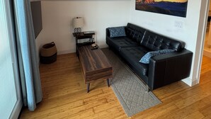 Un salon moderne avec un canapé en cuir noir, une table basse en bois, une lampe et un tableau représentant un paysage de montagne.