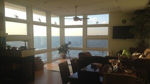 A spacious living area with large windows overlooking the sea, a dining table, and a ceiling fan.