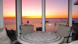 A round table with a tiled top, surrounded by two beige armchairs, overlooking a sunset over the water with boats visible.