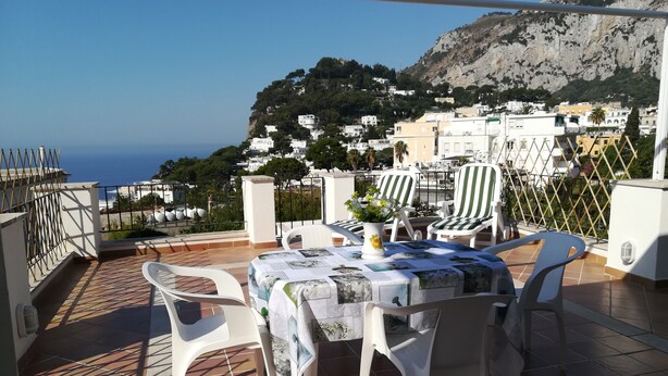 Eine Terrasse mit Tisch und Stühlen, einem Blumenstrauß und Blick auf das Meer sowie Gebäude im Hintergrund.