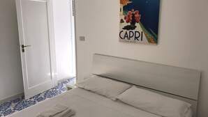 Ein Schlafzimmer mit einem Bett, weißen Kissen und einem gefalteten Handtuch. Eine Tür führt in einen Raum mit blau-weiß gemustertem Boden. An der Wand hängt ein Poster von Capri.