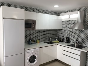 A compact kitchen with white cabinets, a built-in microwave, a stainless steel kettle, and a washing machine.