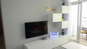 A modern living room with a flat-screen TV displaying 'NETFLIX', a 'WELCOME GUESTS!' sign, and a 'Dream' sign on the wall.