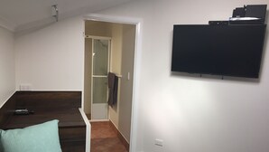 A living room with a dark wood floor, a sofa with patterned cushions, a flat-screen TV mounted on the wall, and a remote control on the floor.