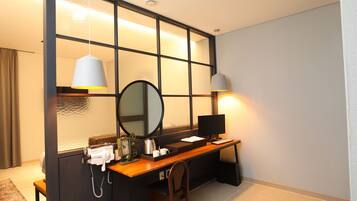 A hotel room with a wooden desk, a chair, a large mirror, a black lamp, and a small table with a coffee pot and cups.
