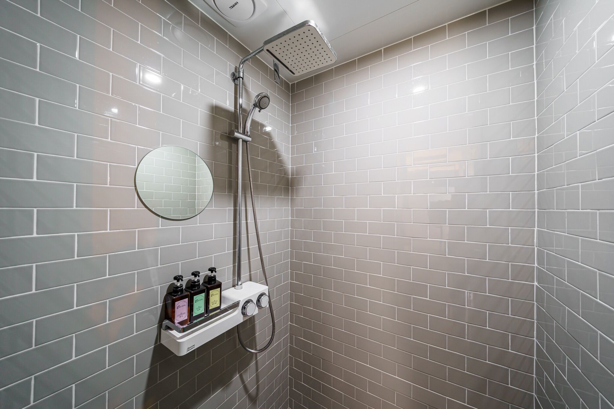 A shower with a handheld showerhead, a wall-mounted mirror, and a soap dispenser.
