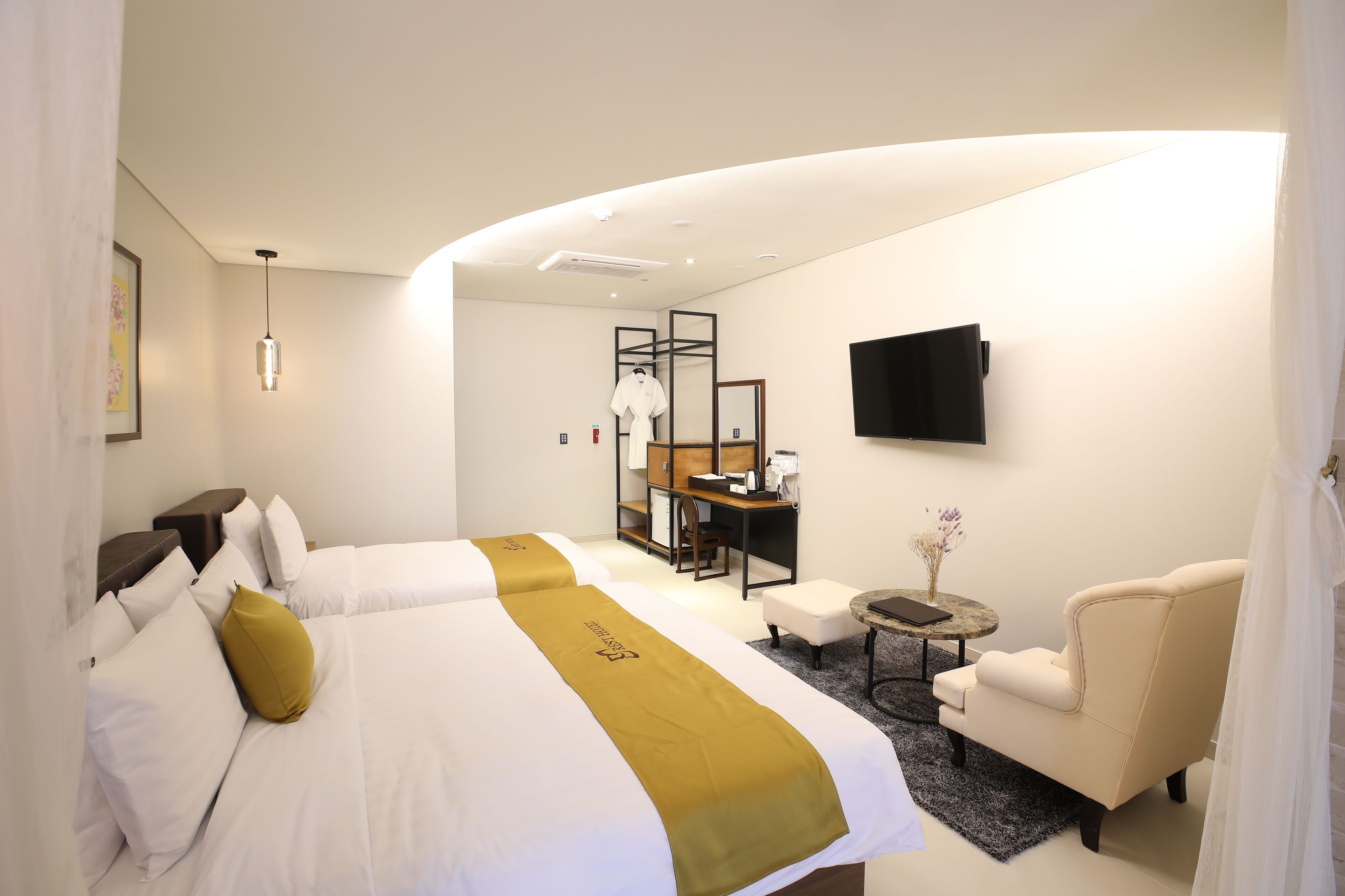 A hotel room with two beds, a desk, a chair, a TV, and a small table with a vase.