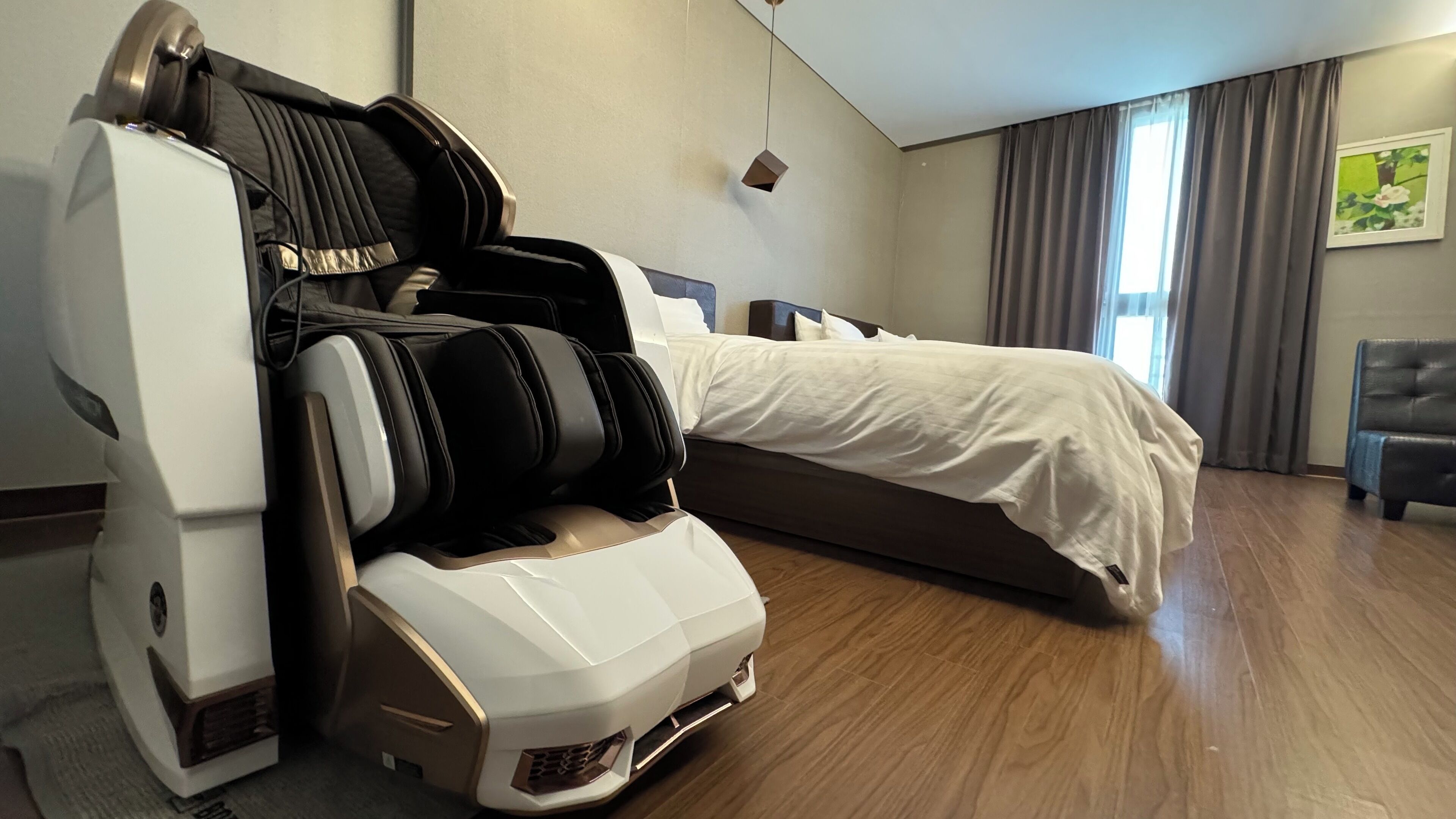 A hotel room with a massage chair, a bed, a bedside table, and a window with curtains.
