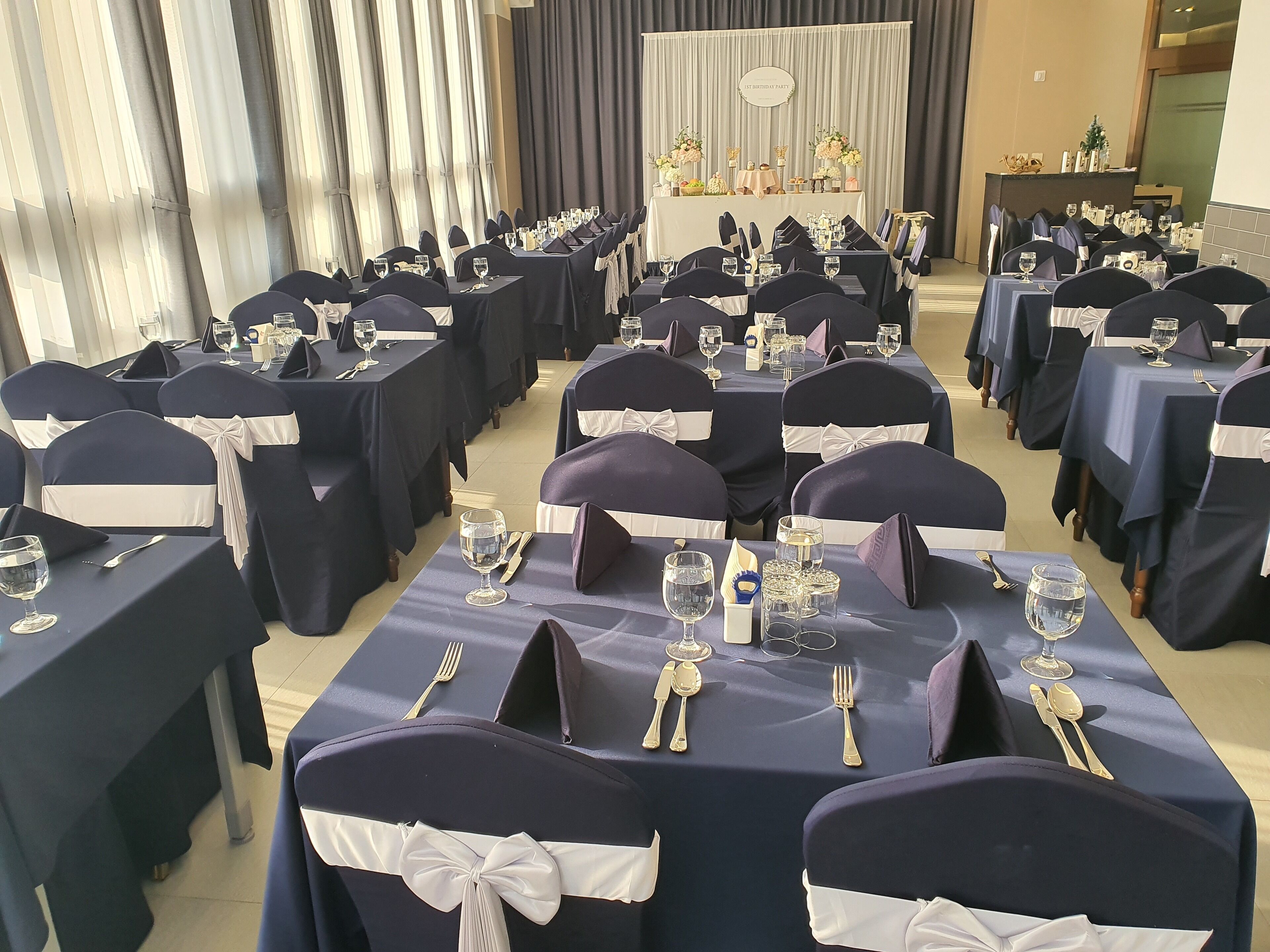A banquet hall with rows of tables set for a formal event, each with a white table runner and dark chairs with white sashes.