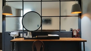 Une pièce comprenant un bureau en bois, un miroir rond, deux lampes suspendues, une chaise et une fenêtre avec un vitrage texturé.