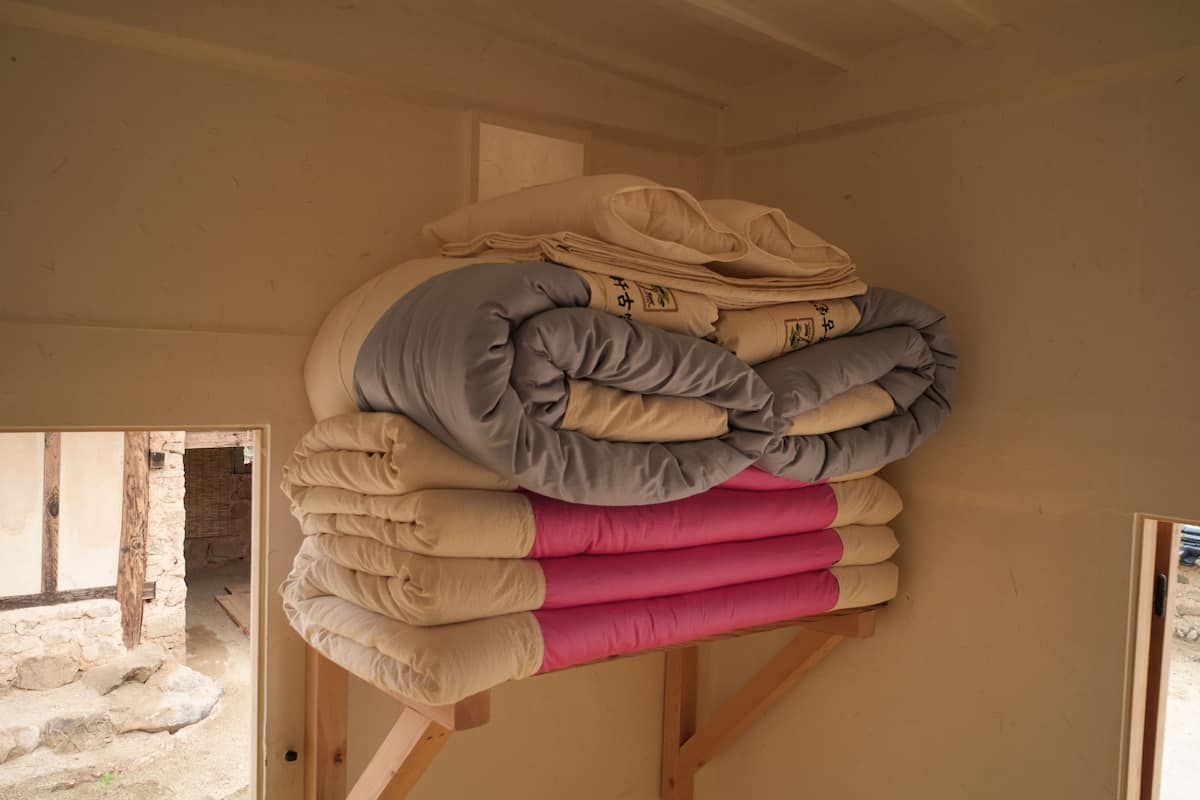 A stack of pillows in various colors on a wooden stand in a room with stone walls and a wooden floor.