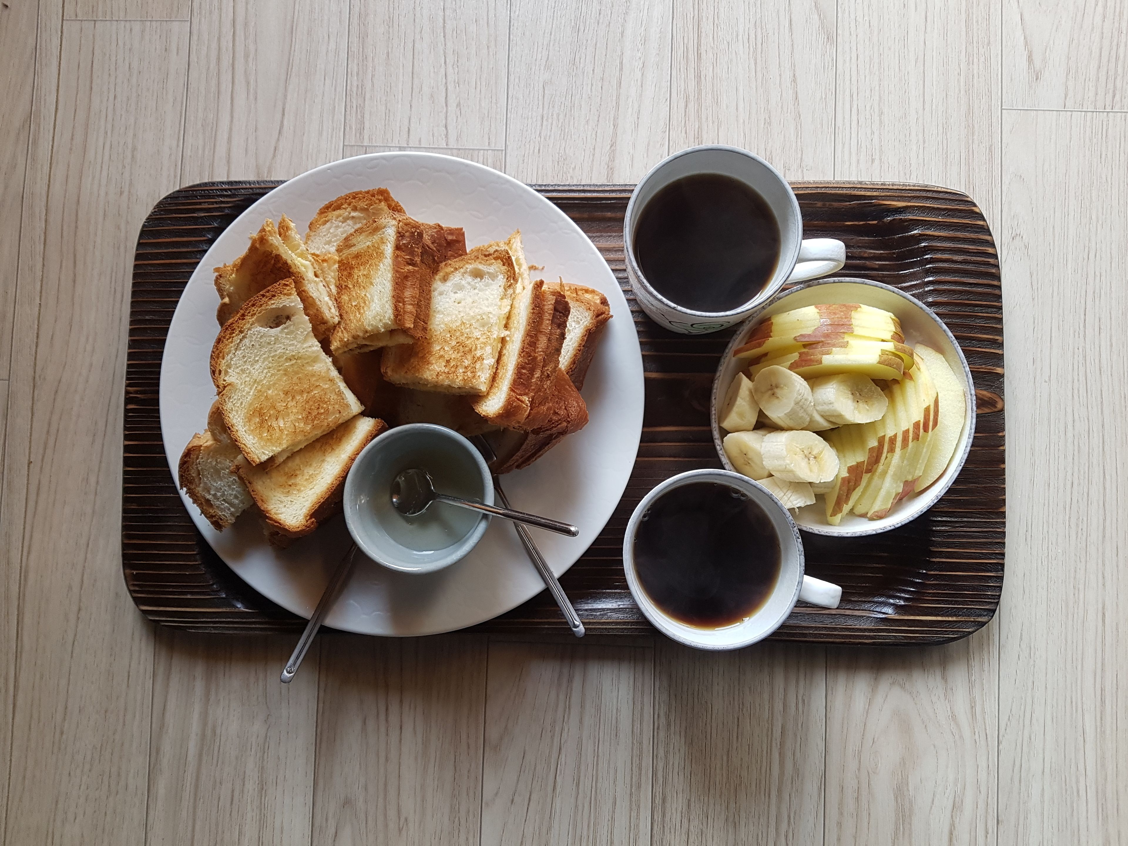 Una bandeja con pan tostado, rodajas de plátano y dos tazas de café sobre una mesa de madera.