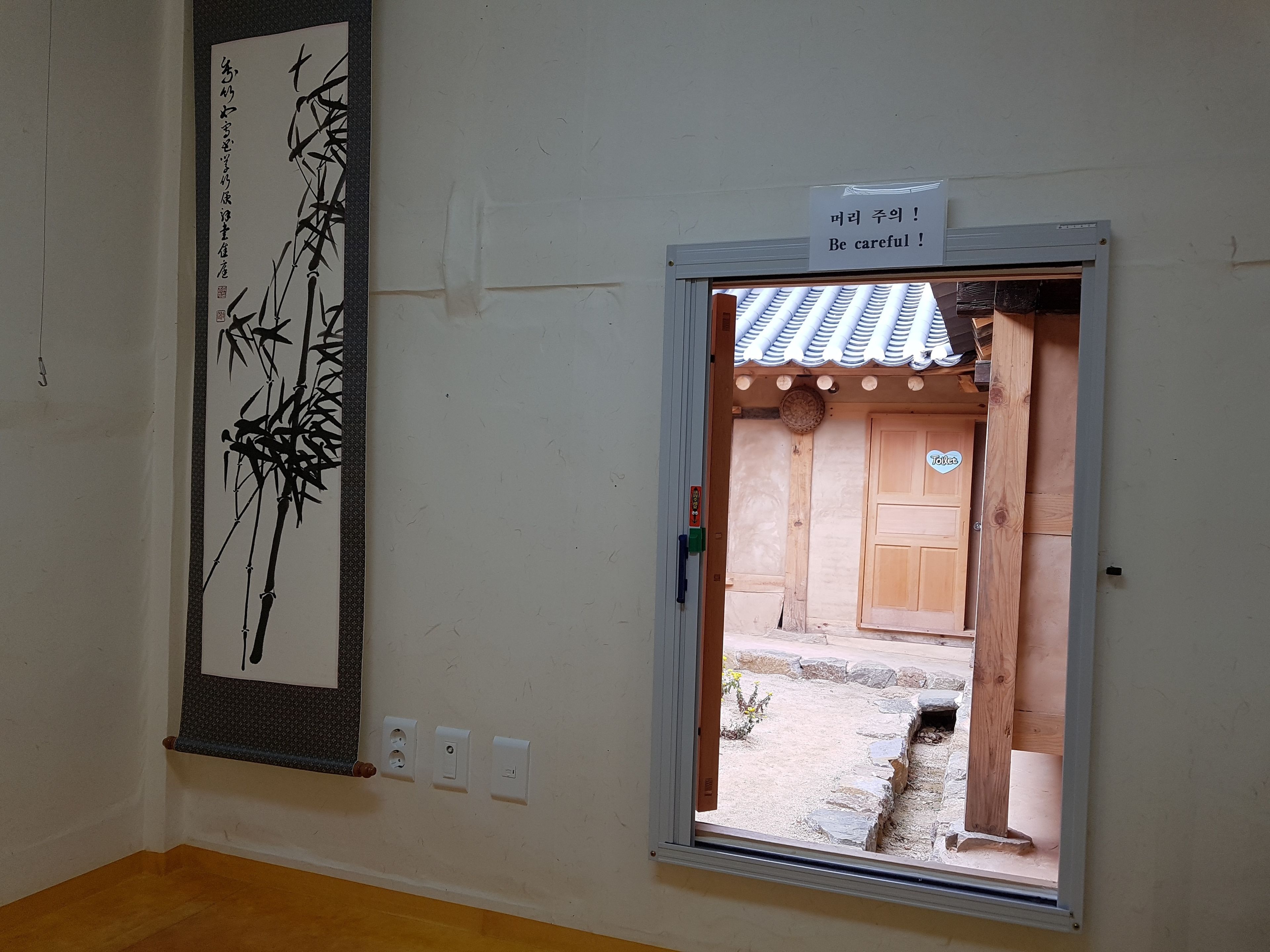 A room with a traditional Japanese calligraphy artwork on the wall, a window with a 'Be careful!' sign, and a wooden door labeled 'Toilet'.