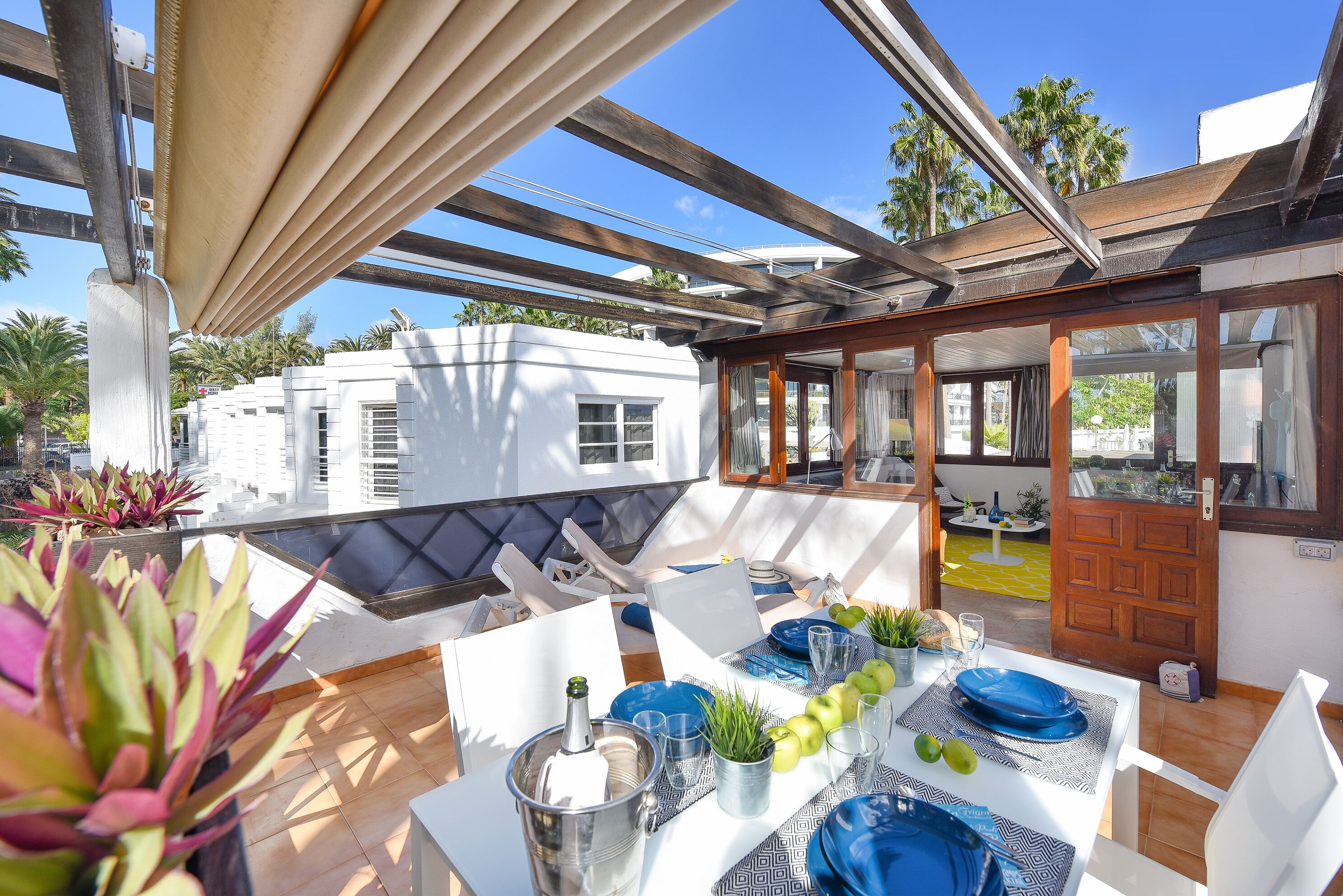 A rooftop terrace with a dining table set for four, surrounded by potted plants and a view of palm trees and buildings.