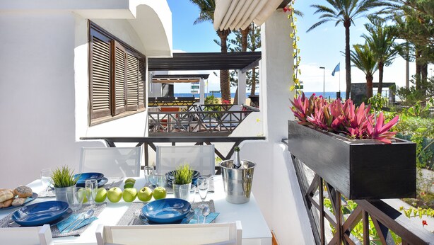 En terrasse med spiseplads, inklusiv et bord med tallerkener, glas og en champagneflaske. Der er plantekasser og udsigt til havet og palmer i baggrunden.
