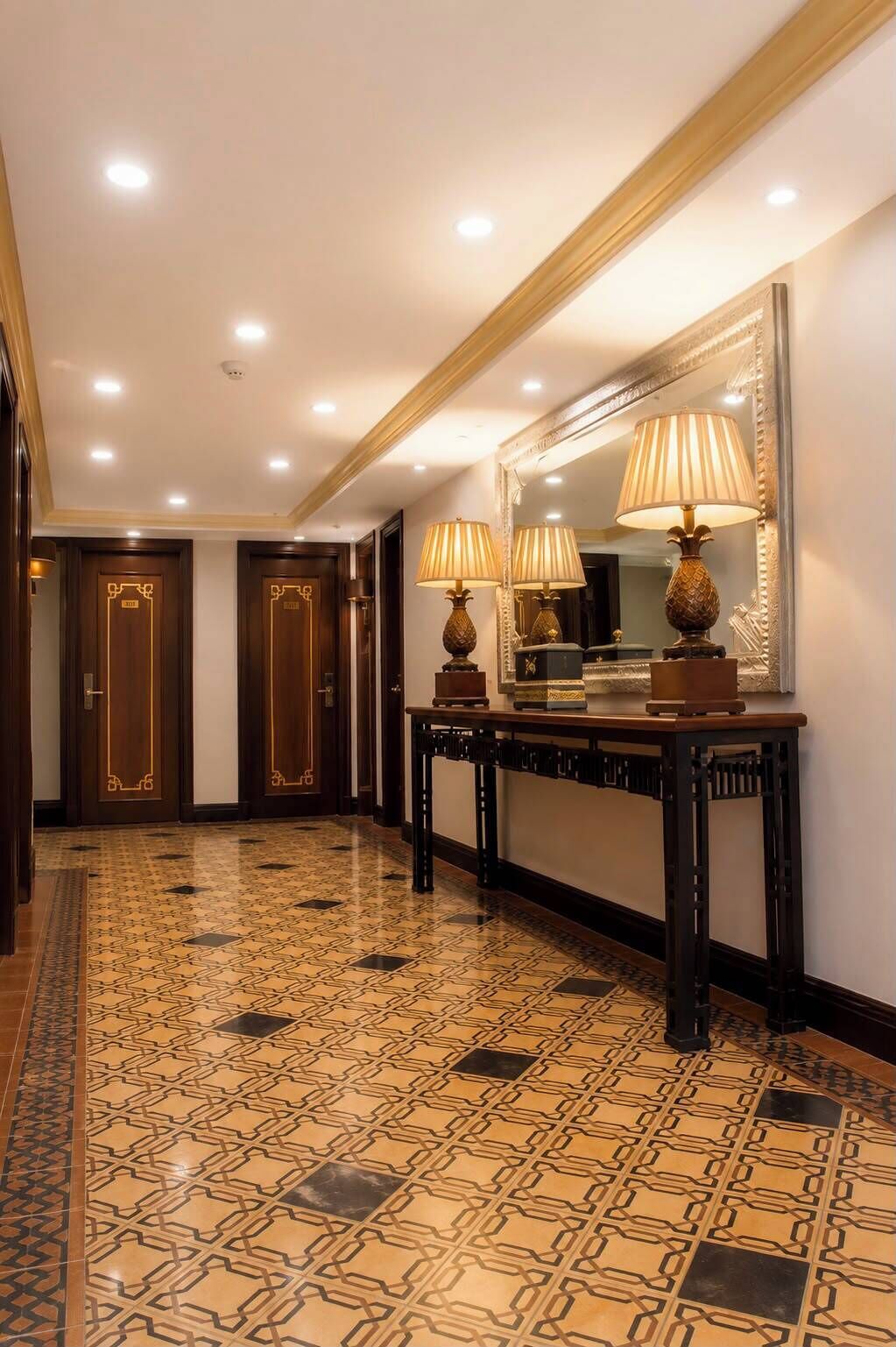 A hotel hallway with patterned flooring, wooden doors, and a console table with lamps and a mirror.