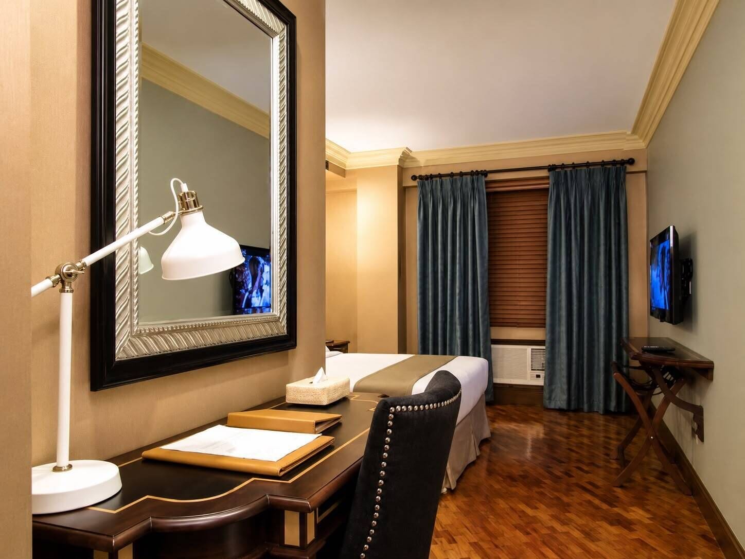 A hotel room with a bed, a desk with a lamp, a television, and a large mirror.