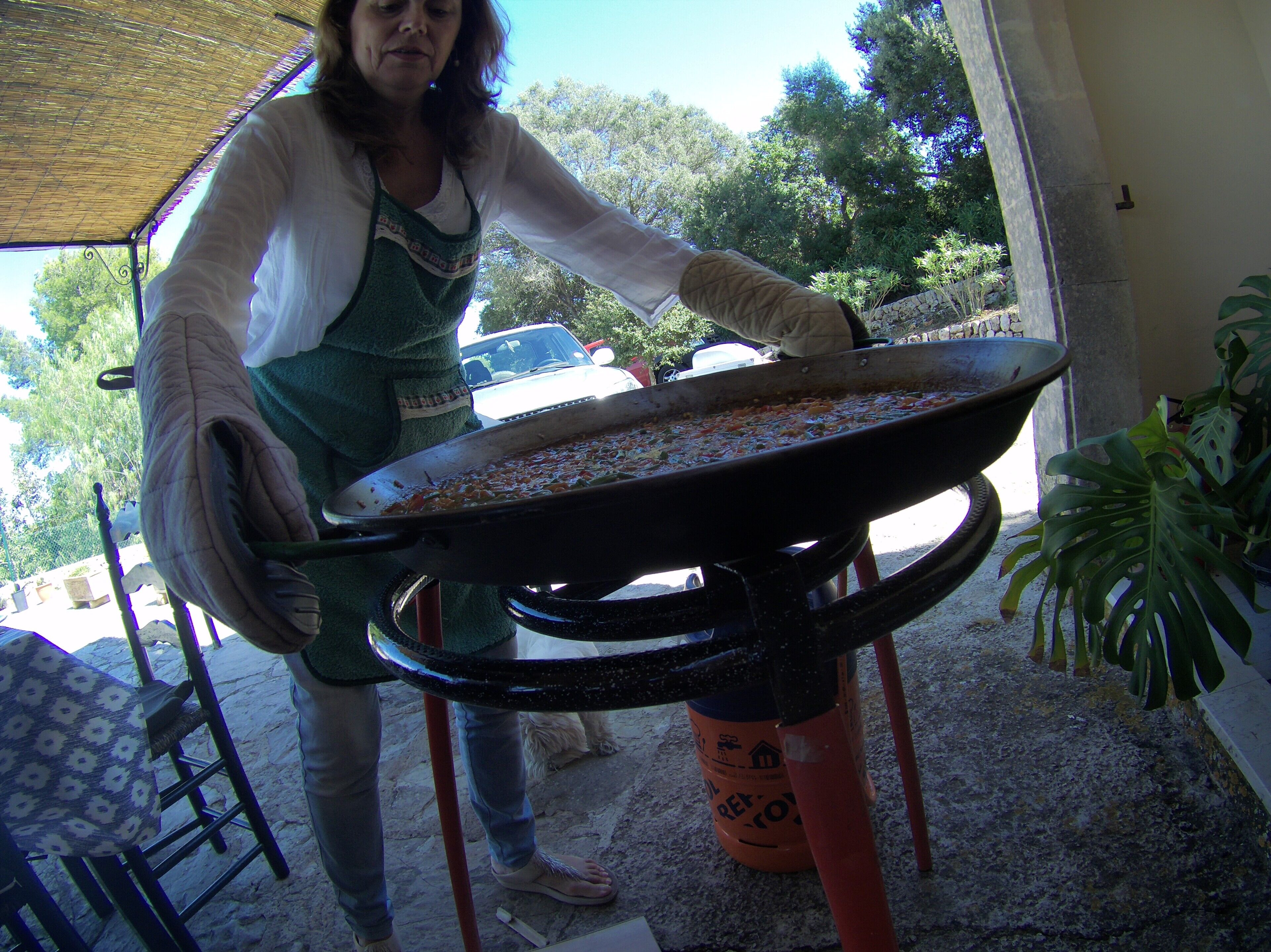A person holding a large pan with food, standing in an outdoor area with a chair and a potted plant nearby.