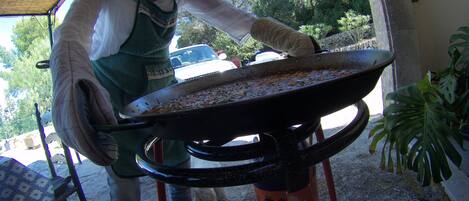 A person holding a large pan with food, standing in an outdoor area with a chair and a potted plant nearby.
