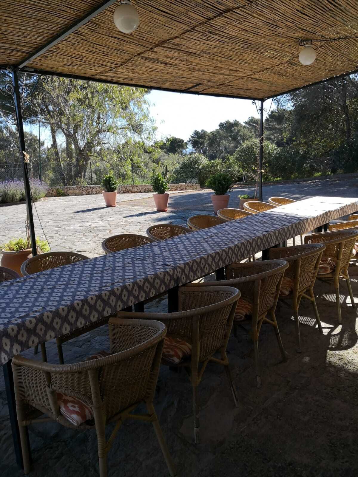 An outdoor dining area with a long table covered with a patterned tablecloth, surrounded by wicker chairs.
