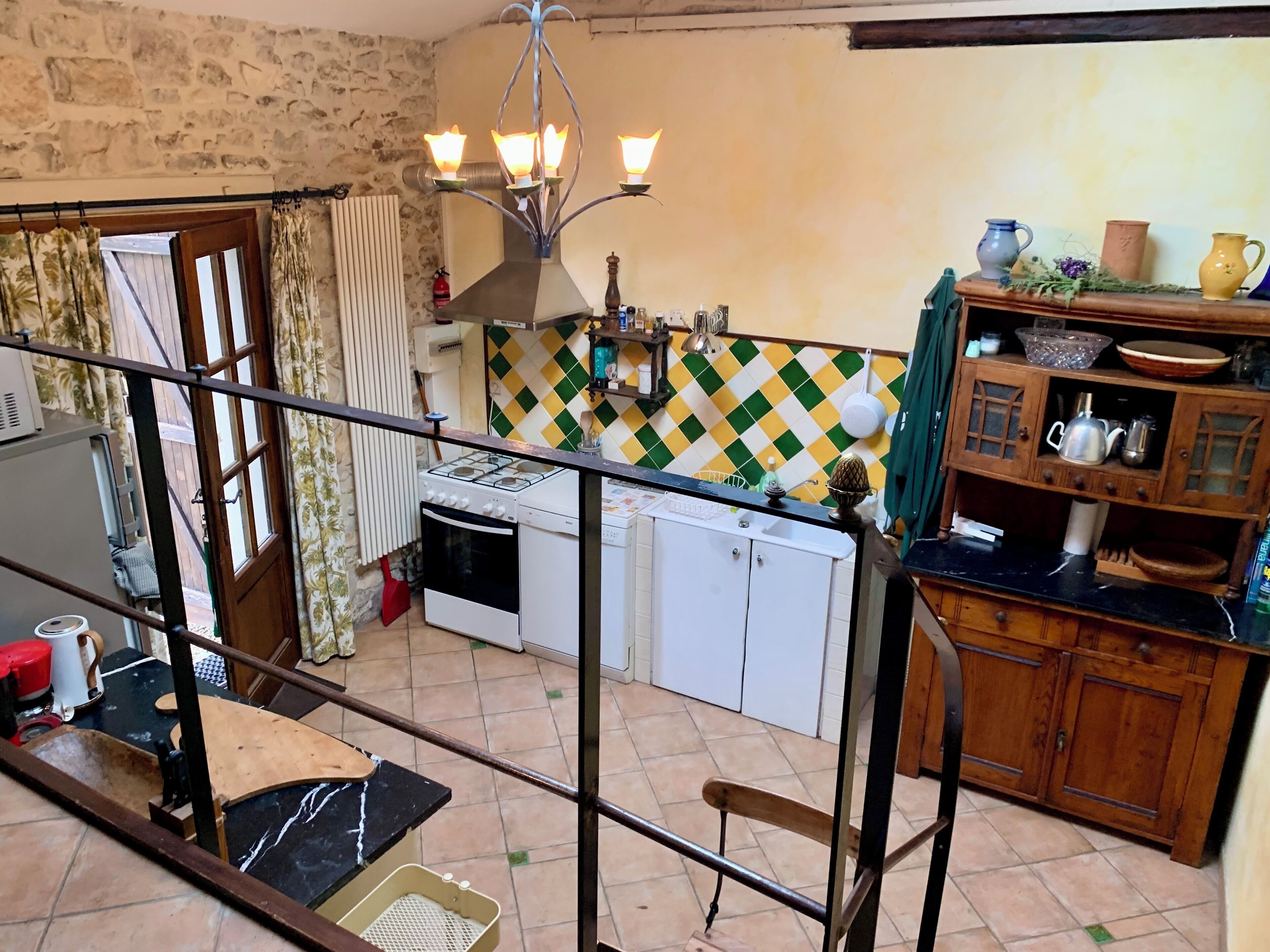 A kitchen with a gas stove, oven, and white cabinets. There is a wooden shelf with kitchenware and a tiled backsplash with a green and yellow pattern.