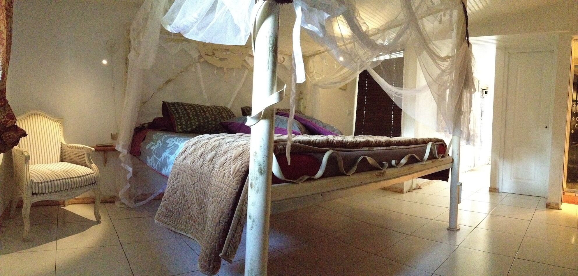 A four-poster canopy bed with a white canopy, a striped armchair, and a bedside table.
