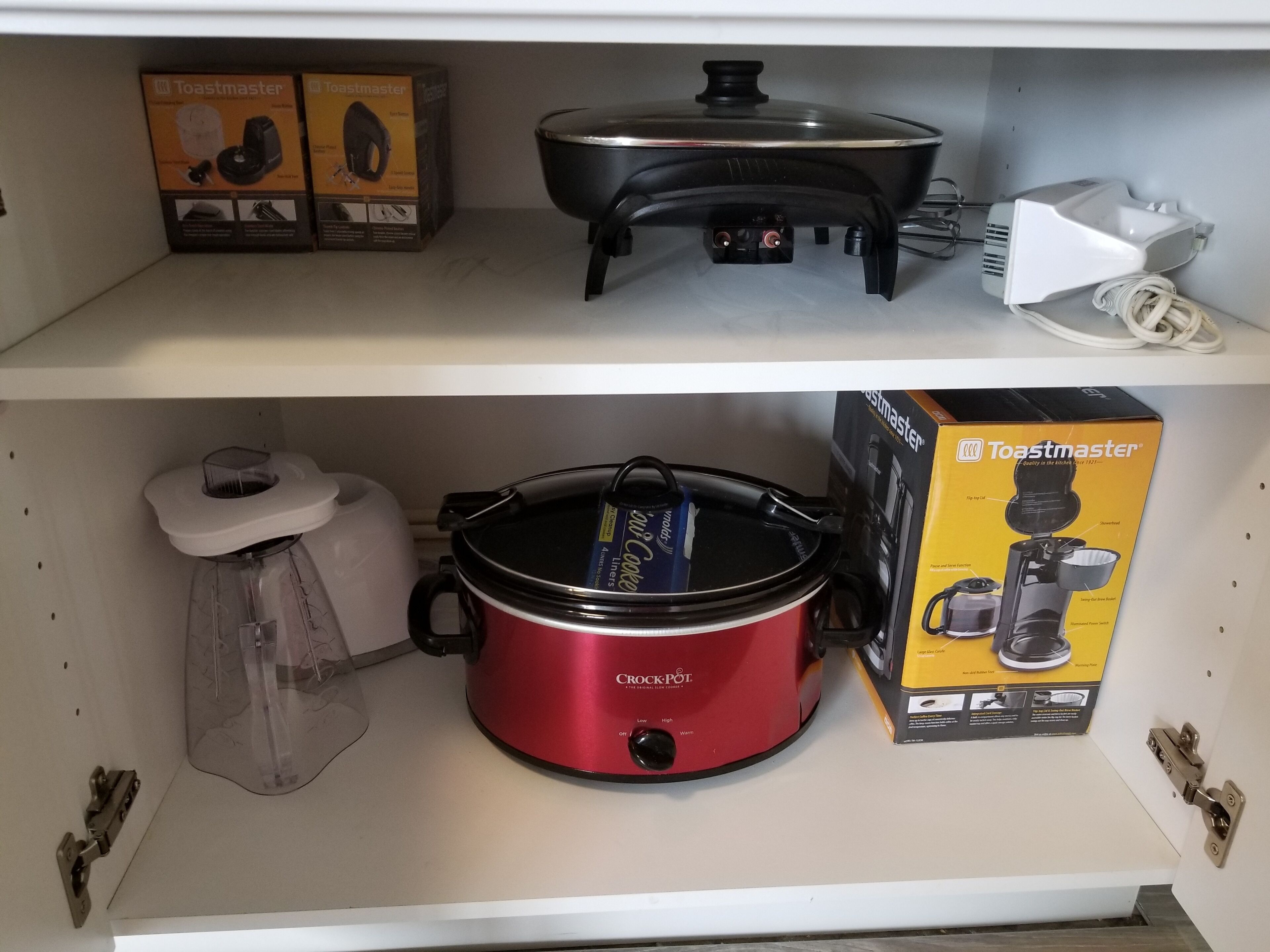 A kitchen shelf with a Toastmaster toaster, a black electric kettle, a red slow cooker, and a white kettle.