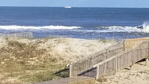 Un paysage côtier avec une promenade en bois menant à la plage, des vagues se brisant sur le rivage et un navire à l’horizon.