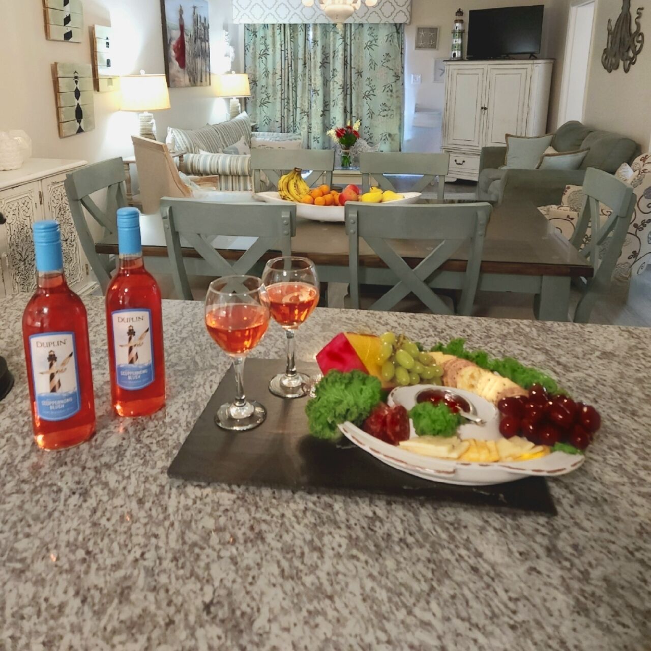 A dining table with a fruit platter, two bottles of red wine, and three wine glasses.