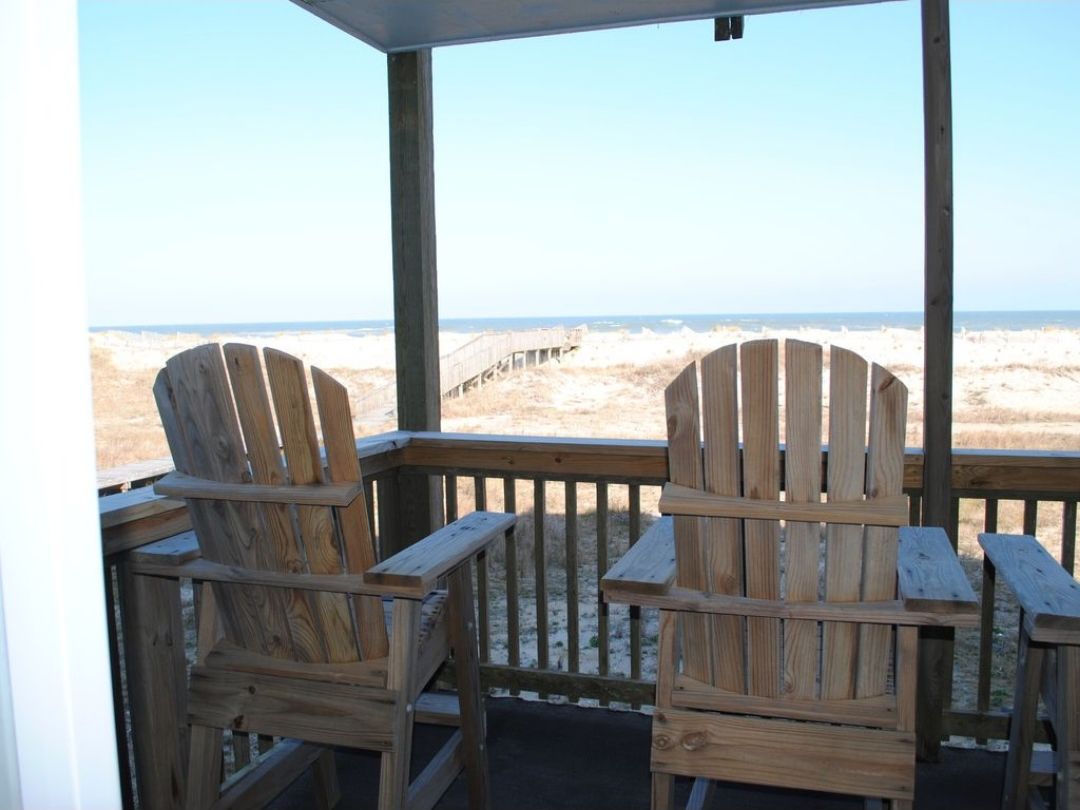 A wooden deck with two Adirondack chairs facing a beach.