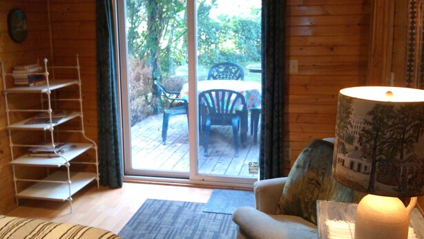 Ein gemütliches Zimmer mit Holzvertäfelung, einer Schiebetür zu einer Terrasse, einem Bücherregal, einem gepolsterten Sessel und einem Tisch mit Stühlen.