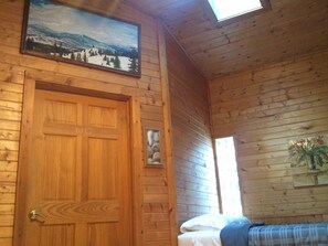 A wooden cabin room with a bed, a door, and a painting of a snowy landscape.