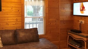 Ein gemütliches Zimmer mit Holzbalkendecke, einem braunen Ledersofa, einem Fernseher und einem Fenster mit Jalousien.