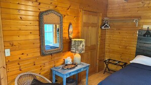 Ein Holzhüttenraum mit einem Bett, einem Korbstuhl, einem Tisch mit Lampe und einem Spiegel an der Wand.