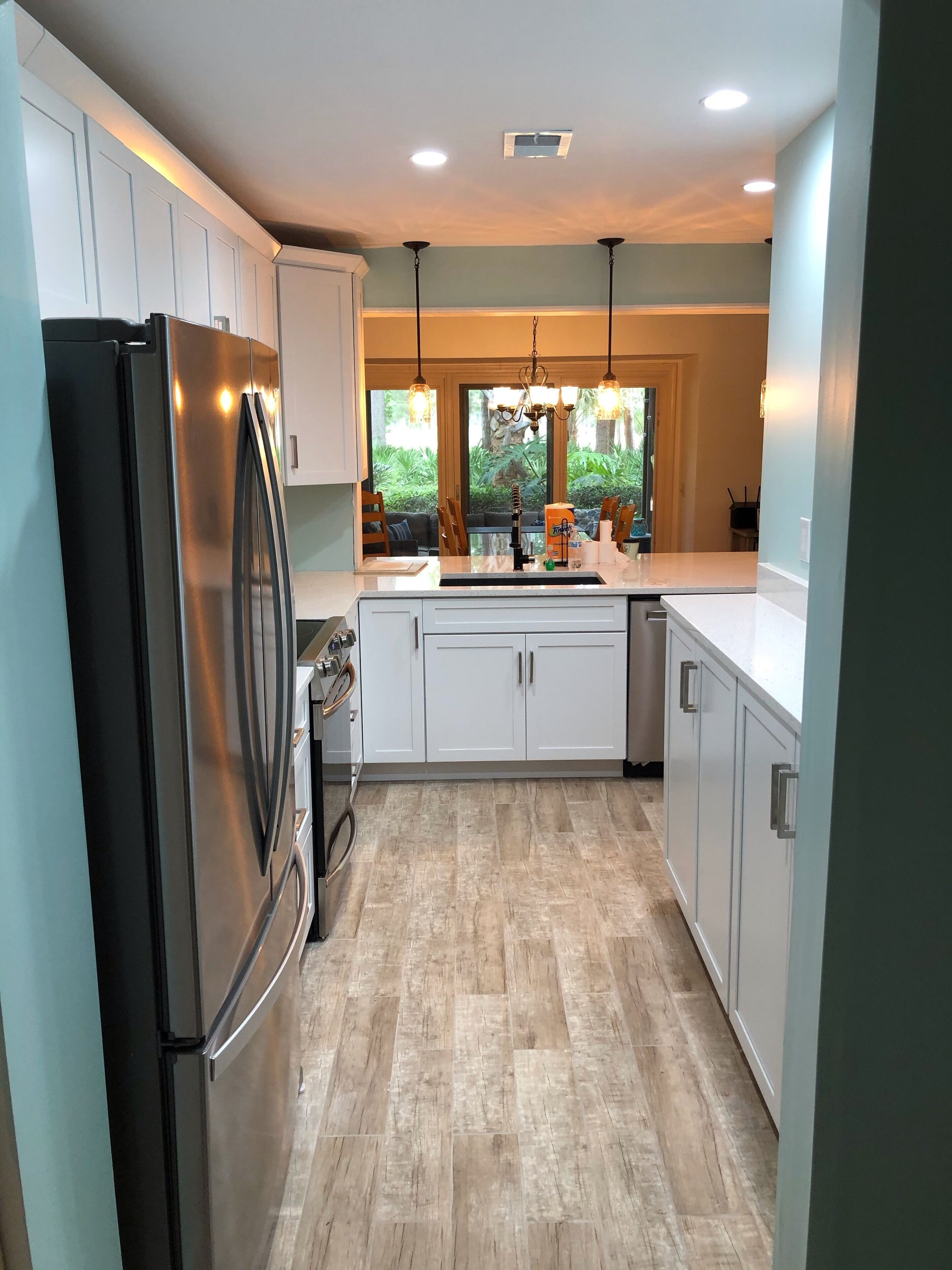 A modern kitchen with white cabinets, a stainless steel refrigerator, and a large window providing a view of greenery outside.