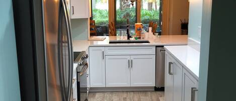 A modern kitchen with white cabinets, a stainless steel refrigerator, and a large window providing a view of greenery outside.
