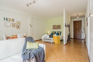 A living room with a sofa, a dining table with chairs, a kitchen area, and a hallway leading to a door.