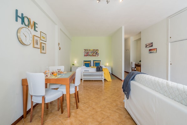 A modern, well-lit room with a dining area, a bed with a blue and yellow bedspread, and a white sofa.