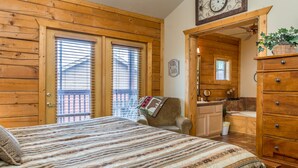 Quarto de cabana de madeira com uma cama, uma cadeira, cômoda e um banheiro visível por uma porta aberta.