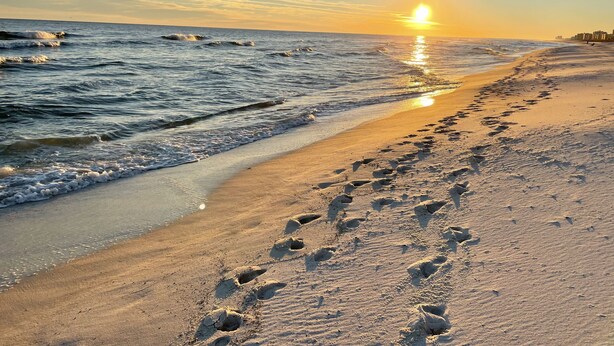 Een strand met voetsporen die naar het water leiden, golven en een zonsondergang.