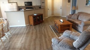 A living room with a grey sofa, a wooden coffee table, and a kitchen area with white cabinets and a refrigerator.