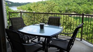 A balcony with a round table and four chairs, overlooking a lake and forested hills.