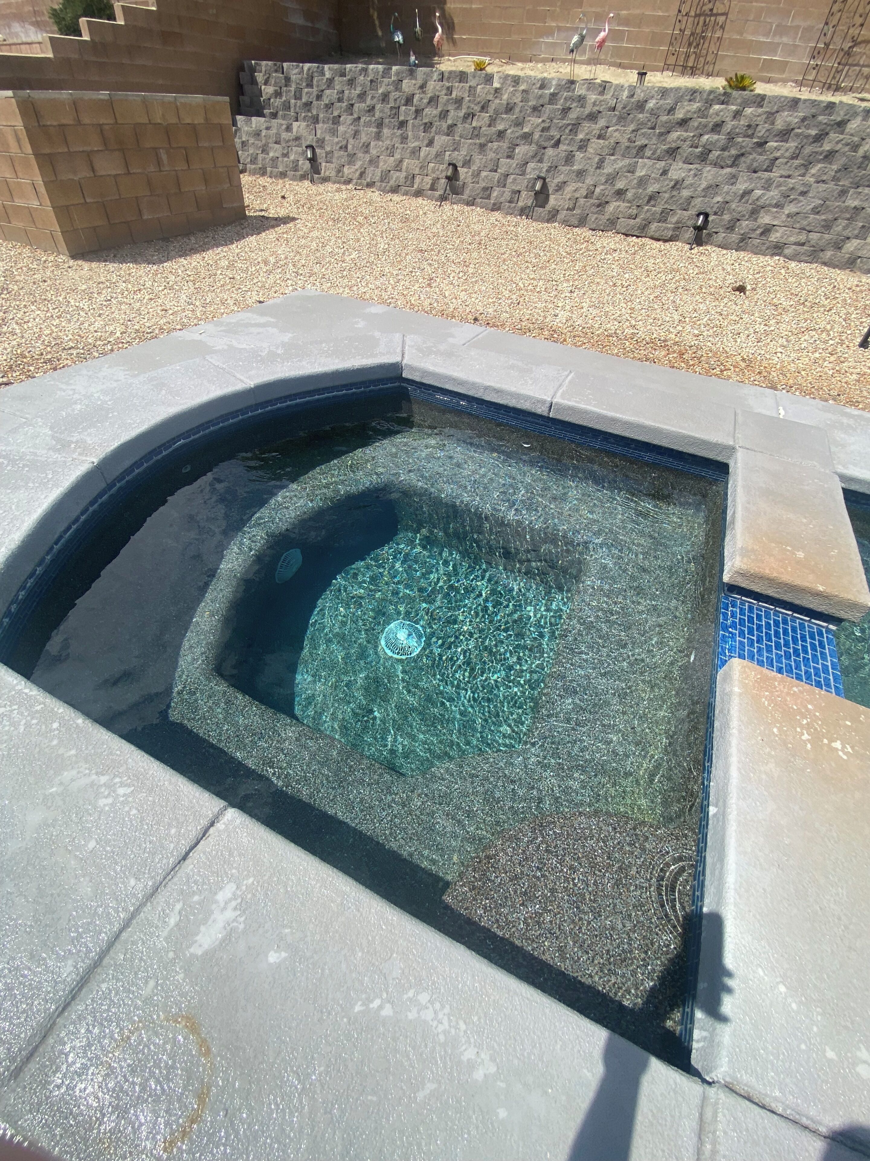 A clear swimming pool with a tiled edge, surrounded by a pebble ground and a brick wall.
